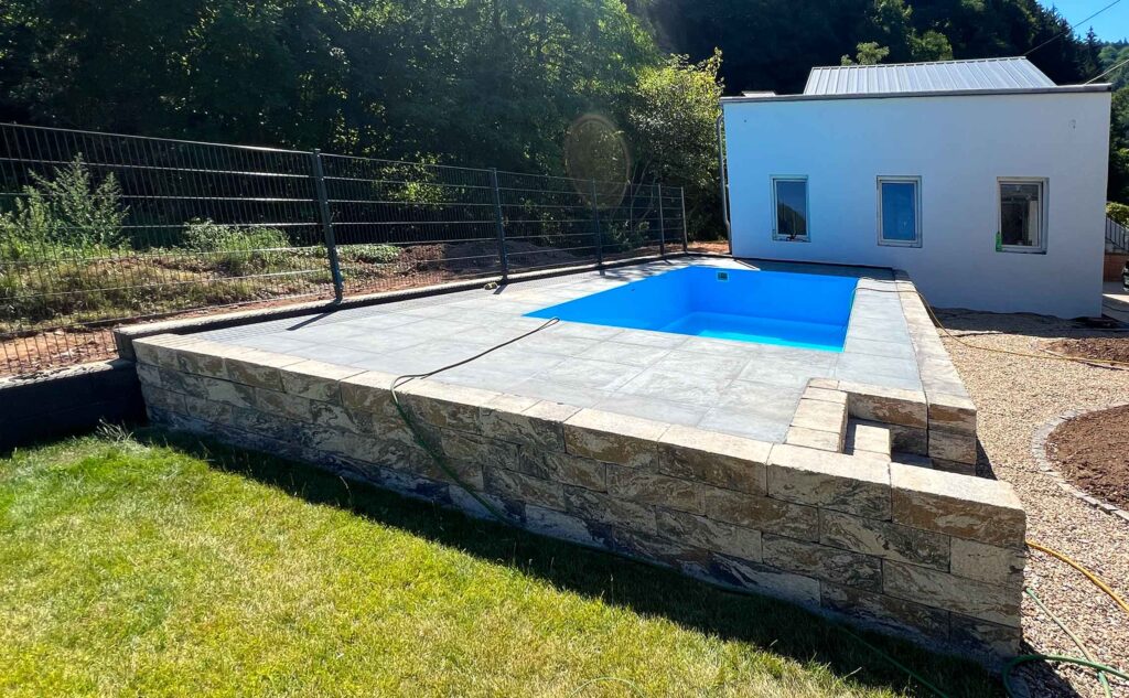 Poolbau eines kleinen blauen Pools in einem Podest mit Terrasse, Mauer und Stufen zum Pool in einem angelegten Garten mit Zaun