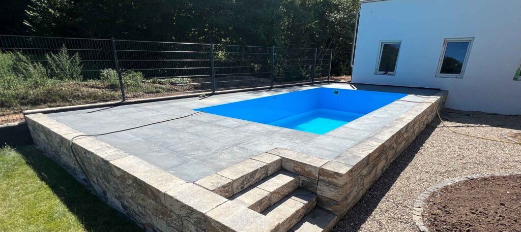 Poolbau eines kleinen blauen Pools in einem Podest mit Terrasse, Mauer und Stufen zum Pool in einem angelegten Garten mit Zaun